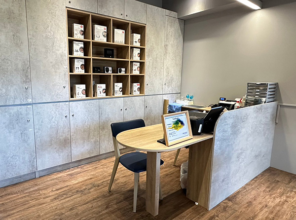 元健助聽器台北內湖店門市內部，灰色系收納櫃與木格展示櫃陳列助聽器產品，前方為木質桌面與藍色座椅的諮詢區，旁有灰色隔板與聽力檢測設備。