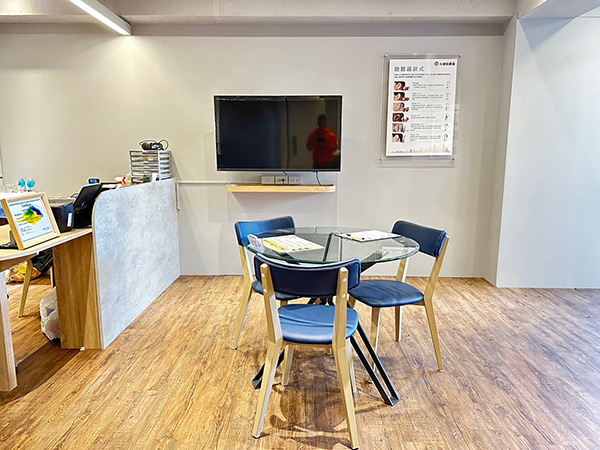 元健助聽器台北內湖店試聽區，玻璃圓桌搭配藍色椅子，牆上掛有電視螢幕與聽力資訊海報，木紋地板明亮整潔。