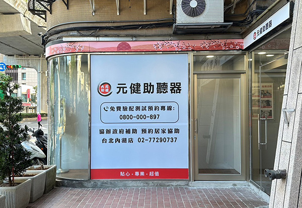 元健助聽器台北內湖店門市外觀,弧形玻璃門面貼有藍白色大型看板,寫著元健助聽器、免費驗配與試戴預約專線0800-000-897、協辦政府補助、預約居家協助,以及台北內湖店電話02-77290737,門口種有綠色植栽。