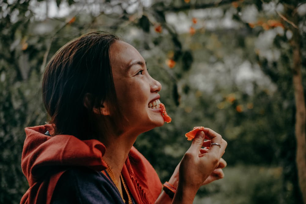 身穿紅色連帽外套的女性在戶外樹林旁開心地笑著吃東西的封面圖，點擊閱讀〈防止聽力損失》你該怎麼吃?〉，了解吃什麼對耳朵聽力最有幫助