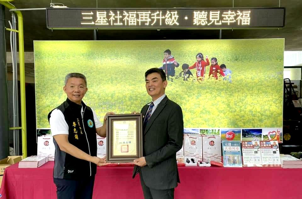 元健天籟列車宜蘭三星鄉活動現場,兩位男士在「三星社福再升級・聽見幸福」跑馬燈與油菜花田背景板前合捧感謝狀合影,桌上陳列助聽器禮盒,點擊閱讀〈元健「天籟列車」停靠三星鄉 免費提供500台助聽器〉