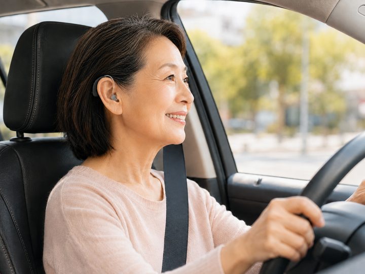 一位熟齡女性繫好安全帶握著方向盤開車，耳上配戴 UP-6EM 助聽器、面帶微笑望向前方，呈現開車出門時能安全聽見周遭聲音與導航提示、出行更安心的情境。