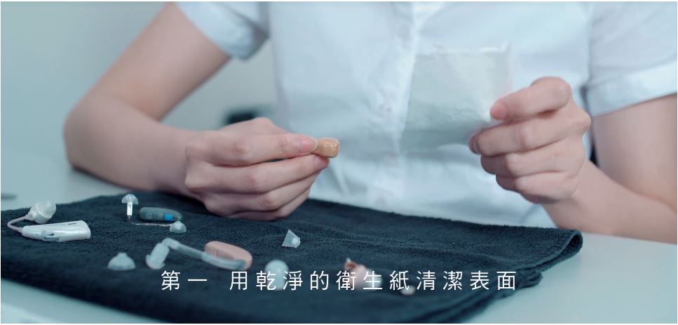 助聽器保養教學影片截圖：身穿白襯衫的人員在黑色毛巾上排放多款助聽器與耳塞配件，字幕寫著「第一 用乾淨的衛生紙清潔表面」，點擊閱讀〈如何保養助聽器？聽力專家教學：清潔助聽器這樣做！讓助聽器多用數年！〉