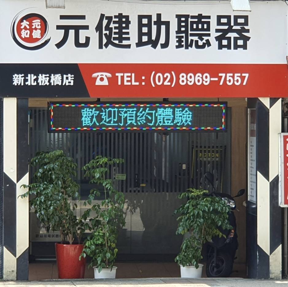元健助聽器新北板橋店門市外觀，白色與紅色招牌印有元健助聽器標誌、新北板橋店與電話(02)8969-7557，門口上方LED跑馬燈顯示歡迎預約體驗，門前擺放大型綠色盆栽