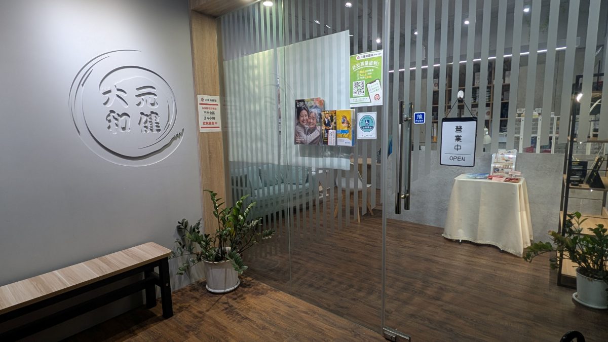 元健助聽器基隆仁愛店門市入口,灰色牆面上有立體的元健助聽器圓形標誌,旁邊為木條隔柵與玻璃門,門上張貼活動海報,地面為木紋地板並擺放木長凳與綠色盆栽。