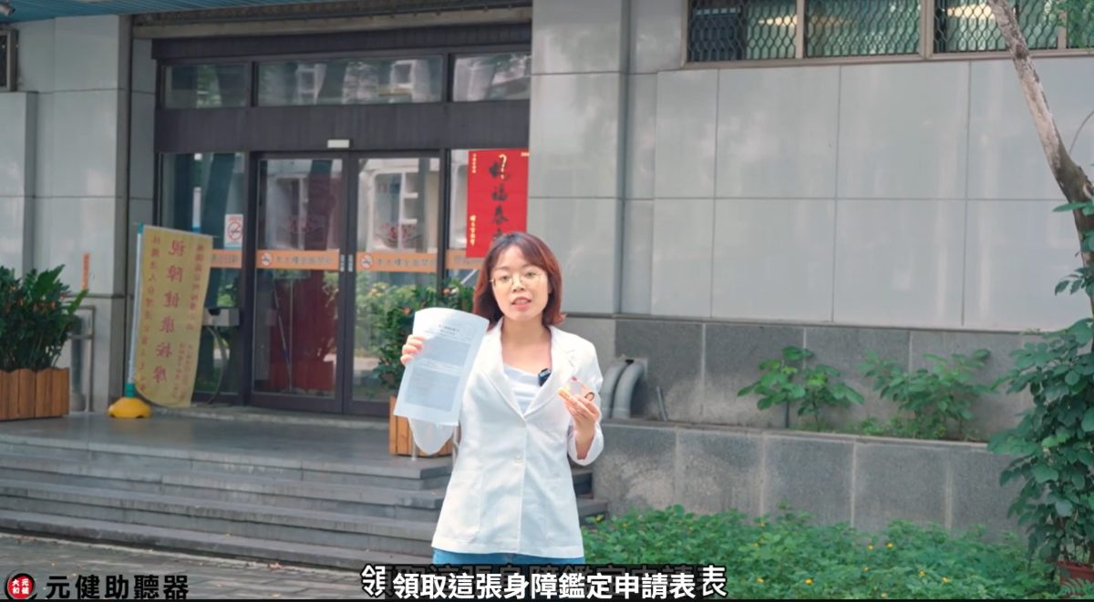 元健助聽器影片截圖：身穿白色外套的女性站在公所大門前手持一張表單說明，字幕寫著「領取這張身障鑑定申請表」，點擊觀看〈115年助聽器補助辦法說明│2026年助聽器政府補助辦法、金額、申請流程 讓元健聽力中心帶您了解！〉