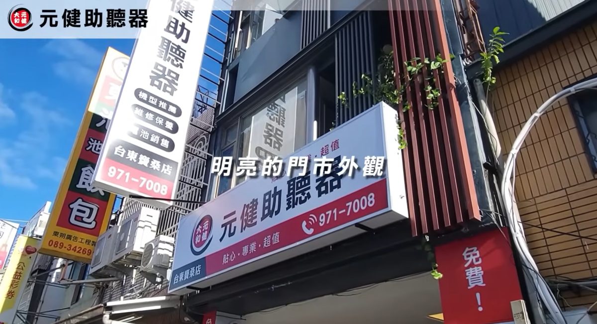 元健助聽器門市外觀影片截圖：藍天下的騎樓店面掛著紅白色元健助聽器招牌與電話971-7008，字幕寫著「明亮的門市外觀」，點擊觀看〈元健助聽器 - 門市印象〉